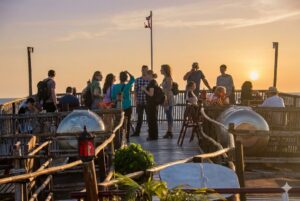 A group of people socializing on the wooden top deck of the Queen Tara riverboat. The scene is bathed in a warm orange sunset over the Tonlé Sap lake, featuring bamboo railings and decorative lanterns.
