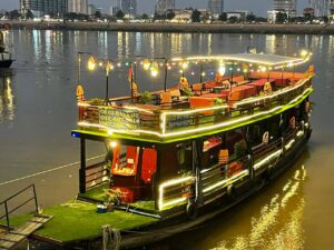 Two-story traditional riverboat illuminated with warm string lights at night, docked on the Mekong River with the Phnom Penh city lights in the background.