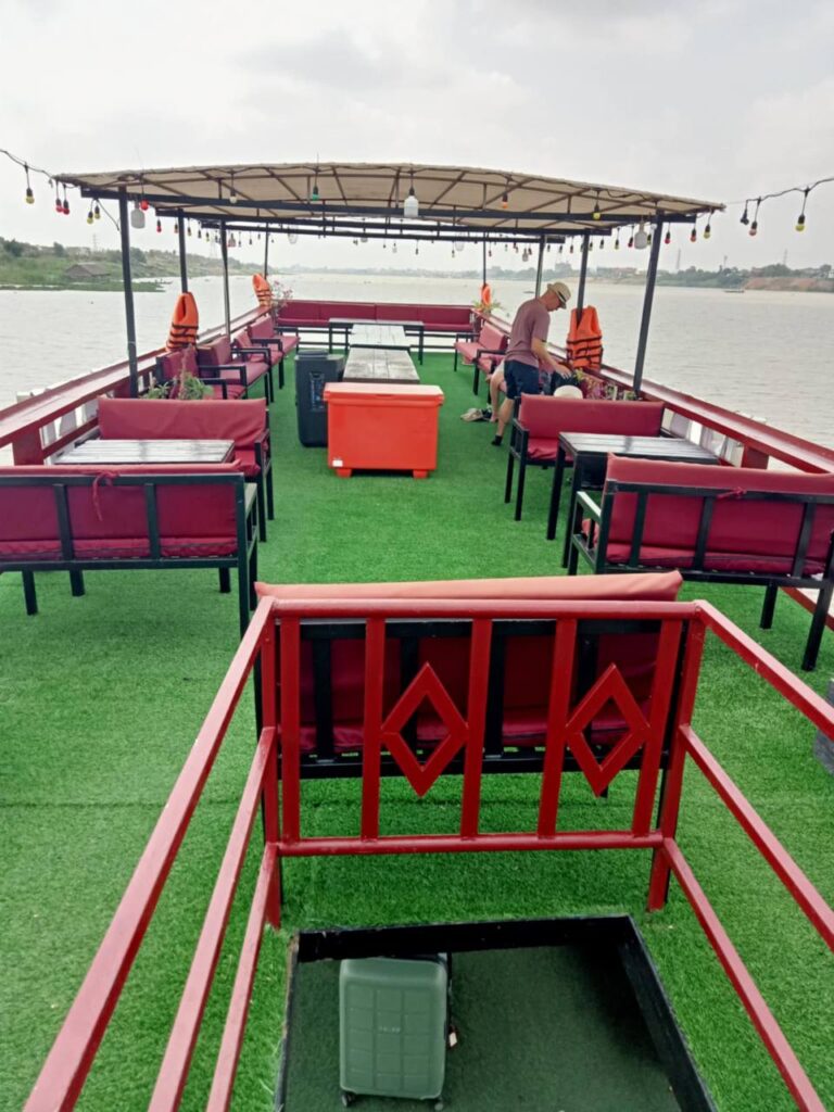 Spacious top deck of a riverboat with red cushioned seating and green turf flooring, cruising the Mekong River between Phnom Penh and Siem Reap.