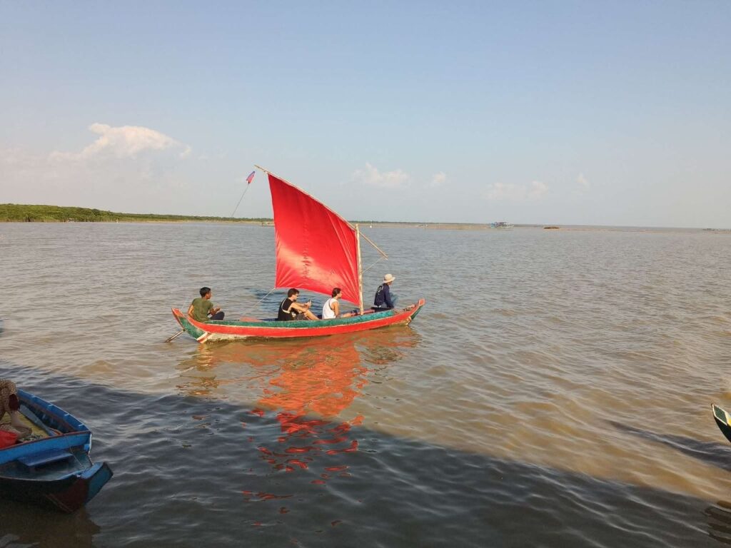 A traditional Khmer wooden sailboat with a vibrant red sail carrying passengers across the calm waters of Tonle Sap Lake near Kompong Phluk, Cambodia.
